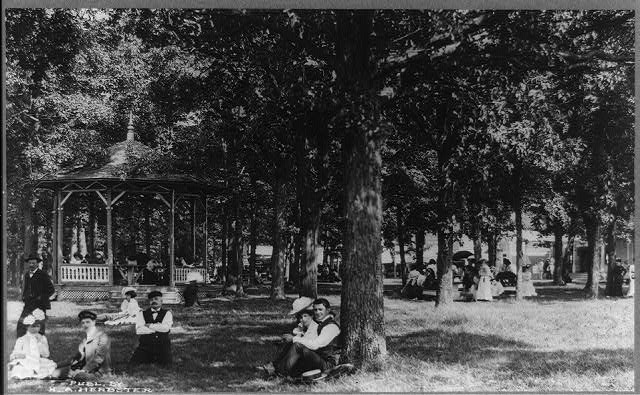 Photograph shows groups of individuals scattered throughout the park either sitting together in the grass or on benches, standing around, or gathered in the gazebo, between 1906-1915